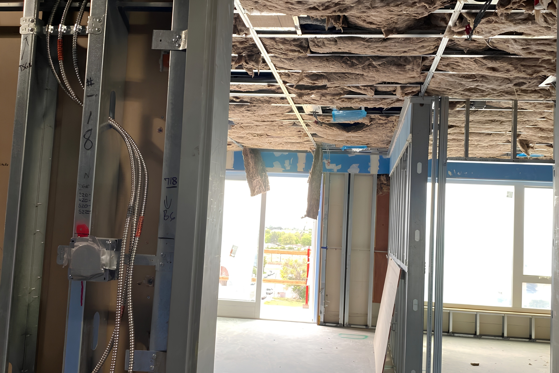 Room under construction with metal stud framing, exposed ceiling insulation, and electrical conduit beside a bright window.